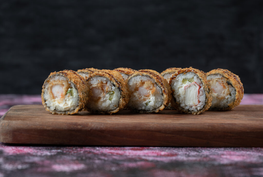 Close-up view of fried hot sushi rolls served on a wooden board, garnished with drizzled sauce, sliced ginger, and a dollop of wasabi on the side.