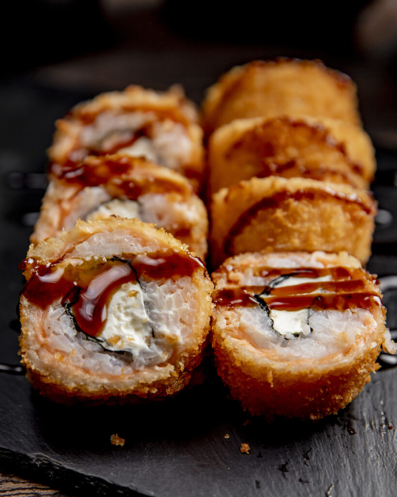 Close-up view of fried hot sushi rolls served on a wooden board, garnished with drizzled sauce, sliced ginger, and a dollop of wasabi on the side.