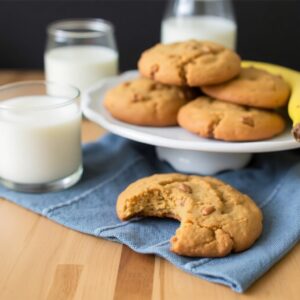 "A plate of freshly baked banana bread cookies with golden edges, showcasing their soft and chewy texture, surrounded by ripe bananas and a glass of milk."