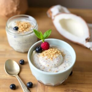 "A glass jar of creamy coconut chia pudding topped with fresh blueberries and shredded coconut, placed on a wooden table."