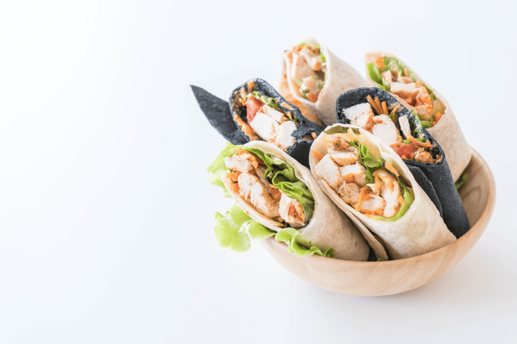 An overhead view of ingredients for shrimp burritos, including tortillas, cooked shrimp, rice, beans, avocado, lettuce, tomatoes, and lime.