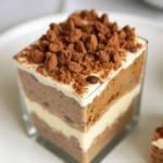 A layered dessert of tiramisu toffee in a glass dish, featuring coffee-soaked ladyfingers, creamy mascarpone, and crunchy toffee bits, dusted with cocoa powder."